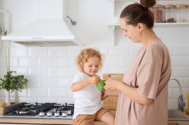 Sevecen anne ve tatlı bebek dalgalı saçlı kızı mutfak masasında oturmuş mutlu aile anları boyunca birlikte sıcak sütün keyfini çıkarıyorlar.