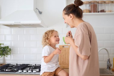 Mutlu anne kahveyi yudumluyor ve sevimli çocuğuyla eğleniyor sıcacık mutfakta sağlıklı içkinin tadını çıkarıyor ve sabahları çay içerken gülücüklerini paylaşıyor.