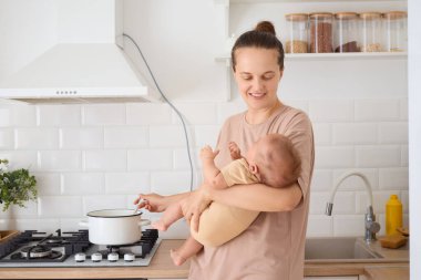 Gülümseyen kahverengi saçlı anne, bebeğini taşırken yemek pişirirken anneliği ve ev işlerini evlerinin sıcaklığında özen ve sevgiyle dengeliyor.