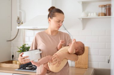 Çok yorulmuş anne mutfakta bebeğini kucağında tutarak sabah rutin yemeklerini yapıyor ve annelik isteğinden bitkin düşmesine rağmen bebeğini taşıyor.
