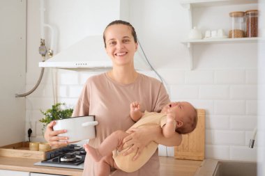 Neşeli anne oğlunu taşırken yemek pişirirken kameraya gülümserken ailesine sevgiyle bakar akşam yemeğini hazırlarken çocuğunu kucağında tutar ve aile işlerinin keyfini çıkarır.