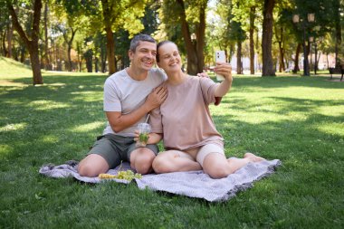 Happy couple picnicking in countryside sitting on blanket enjoying happy time together outdoors laughing and taking selfie with their smartphone embracing romantic moment capturing joyful day clipart