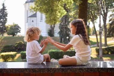 İki kız kardeş parkta oturuyor sarışın bebek dalgalı saçlı kız ablasıyla dışarıda ellerle oynuyor.