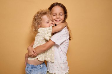 İki mutlu kız kardeş kucaklaşır kollarını. Heyecan ve sevgi gösterirler, neşe saçar ve destek olurlar. Bej arka planda kapalı bir yerde yan yana dururlar.