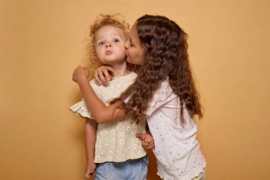 Sevimli kardeşler, sevgi ve şefkat gösteren bir öpücük için dudaklarını şaklabanca büzerler. Mutluluk ve sevgi dolu komik yüzler yaparlar. Bej arka planda kapalı bir yerde dururlar.