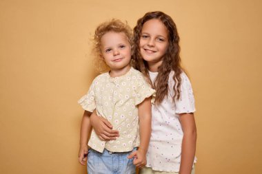 Biri dalgalı sarı saçlı, diğeri kahverengi saçlı, gülümseyen, neşe saçan iki neşeli çocuk ve bej arka planda kapalı yerde duran dostça bir sıcaklık.