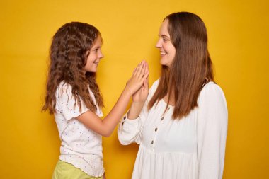 Anne ve kızı, sarı arka planda izole edilmiş neşeli tebessümlerle birbirlerini selamlıyorlar. Sevinç ve dostluğu ifade ediyorlar. Elleriyle beşlik hareketler yapıyorlar.