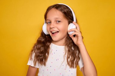 Dalgalı kahverengi saçlı, beyaz tişörtlü, kulaklıkla MP3 dinleyen neşeli bir kız sarı arka planda izole bir şekilde dururken gülümsüyor.