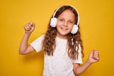 Kahverengi saçlı, kablosuz kulaklıklı, en sevdiği müzik listesini veya podcast dansını dinleyen ve sarı arka planda müziğe kendini kaptırmış gülümseyen kız öğrenci.