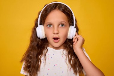 Kahverengi dalgalı saçlı, beyaz tişörtlü, sarı arka planda izole bir şekilde poz veren öğrenci inanılmaz podcast veya kulaklıkla yeni bir şarkı dinlerken hayrete düştü.