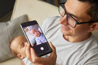 Gözlüklü baba, evde cep telefonuyla fotoğraf çektirirken sevimli yeni doğmuş kızını ya da oğlunu elinde tutuyor ve çocuk odasında güzel bir anı yaratıyor.