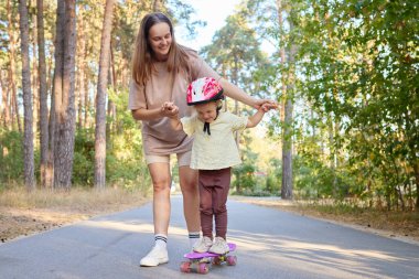Gülümseyen neşeli kadın, eğlenceli ve eğlenceli bir anda, parkta sıcak havanın ve temiz havanın tadını çıkarırken, küçük çocuğuna para tahtasının üzerinde dengede durmasında yardım ediyor.