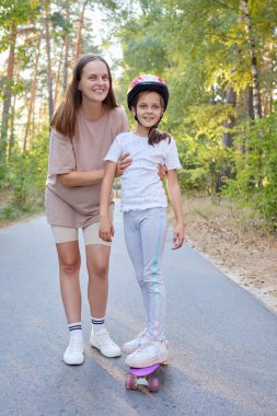 Genç anne kızıyla oynuyor. Yaz parkında kaykayla gezen kızı itiyor. Koruyucu kasklı çocuk, annesiyle spor aktivitelerinin keyfini çıkarıyor.