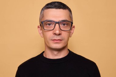 Siyah kazak giyen ve gözlüklü ciddi yakışıklı bir adam kameraya bakıyor sakin bir görünüm ve bej arka planda izole edilmiş odaklanmış kararlılık gösteriyor.