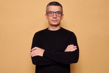 Siyah kazak giyen ve gözlüklü başarılı bir adam kameraya kendinden emin bir şekilde bakıyor. Kollarını çapraz tutuyor. Bej arka planda özlemlerini ve motivasyonlarını gösteriyor.