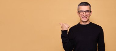 Siyah kazaklı, gözlüklü, neşeli kollu, solu işaret eden mutlu beyaz adam bej arka planda izole edilmiş reklam için fotokopi alanı gösteriyor.
