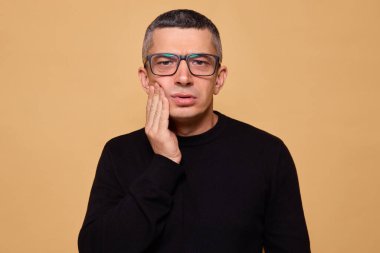 Siyah paçavra kazak giyen üzgün bir adam ve yanağına dokunurken yüzünü buruşturan gözlüklü diş ağrısı çeken bir adamın diş sorunu ve bej arka planda izole edilmiş hassasiyeti var.