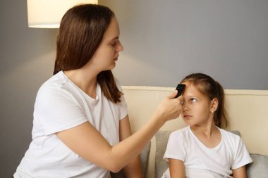 Endişeli bir anne, kızının ateşini dijital termometreyle ölçerken küçük kız kanepede dinleniyor gripten rahatsız oluyor ve evde hasta yatarken bakım arıyor.