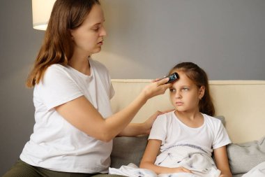 Ebeveyn gribi tedavisi. Tıbbi ateş kontrolü. Hasta genç kız yüksek ateşle kanepede otururken ilgili anne ateşini ölçmek için dijital termometre kullanıyor.
