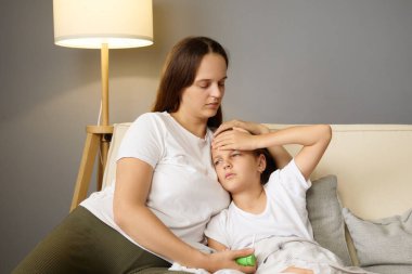 Şefkatli bir anne, hasta kızını kanepesinde kucaklıyor. Ateşli bir kadın var mı diye alnına bakıyor. Evdeki ağrılı hastalığı sırasında onu rahatlatıyor ve ona bakıyor.