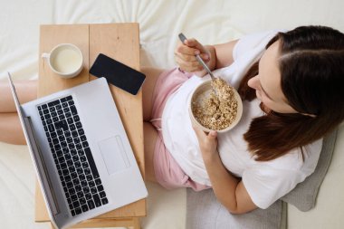 Tanınmayan bir kadın sabah laptopu izlerken ve rahat bir yatak odasında internet üzerinde çalışırken tabakta lezzetli yulaf lapası yiyor.