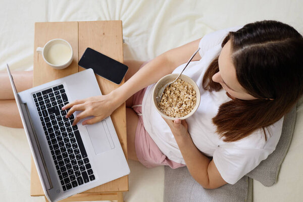 Вид сверху на женщину, завтракающую полезной овсянкой, изучающую онлайн и сидящую на ноутбуке в удобной спальне с ноутбуком и просматривающую Интернет