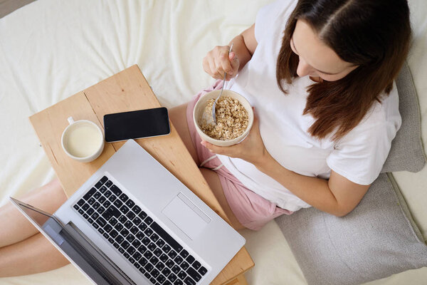 Вид сверху женщины, которая учится, ест вкусную кашу на тарелке и работает на ноутбуке, наслаждаясь социальными сетями в уютной спальне с удобной кроватью и ноутбуком