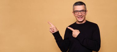 Kazak giymiş mutlu bir erkek, kolunu kaldırmış, boş bir yer gösteriyor, gülümsüyor, dişlek gülümsüyor, kameraya bakıyor, harika hissediyor, reklam alanını bej arka planda yalnız duruyor.