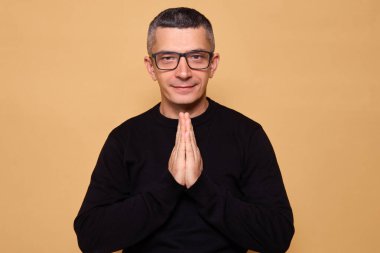 Hopeful man wearing jumper with palms together smiling while asking for forgiveness expressing gratitude feeling positive despite difficulties standing indoor isolated over beige background clipart