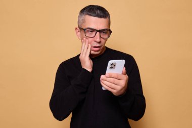Şok olmuş, gözlüklü ve kazaklı bir adam. Akıllı telefona bakıyor, korkmuş ve üzgün. Sosyal medyada bej arka planda izole bir şekilde duran teknolojiyle bağlantılı.
