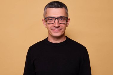 Neşeli pozitif adam gözlük takıyor ve siyah kazak giyiyor bej arka planda izole edilmiş mutlu bir ifadeyle mutlu ve enerji dolu bir şekilde gülümsüyor.