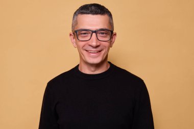 Gözlüklü, siyah paçavra giyen mutlu bir adam bej arka planda izole edilmiş bir şekilde duruyor büyüleyici bir ifadeyle gülümsüyor mutlu bir şekilde kameraya pozitif yüzüyle bakıyor.