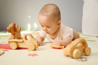 Beyaz tulumlu mutlu çocuk yerde tek başına yatıp ahşap arabalarla oynuyor. Oturma odasındaki renkli paspasın üzerindeki çevre dostu oyuncaklarla göbek zamanının tadını çıkarıyor.
