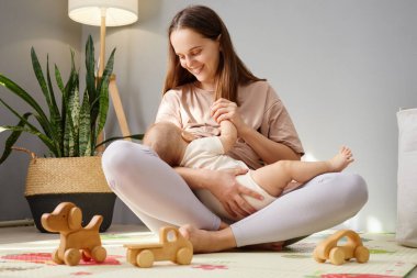 Kahverengi saçlı kadın oturma odasında oturmuş bebeğini sevgiyle emziriyor. Çocuğunu kucaklıyor. Masum çocuğuna sevgi ve sevgiyle bakıyor.