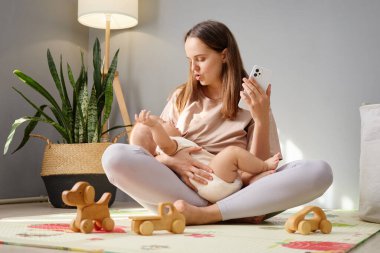 Genç anne, yeni doğmuş bebeğini emzirirken telefonunda ebeveyn tavsiyesi ararken akıllı telefonunu evde ağlayan bebeği sakinleştirmek için kullanıyor.