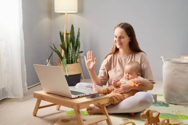 Neşeli kadın doğum izni sırasında evde bilgisayar kullanarak anne şefkati gösteriyor. Dengeli iş ve aile hayatı video araması yapıyor. Selamlaşma işareti yapıyor.