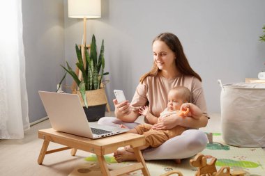 Çalışkan bir iş kadını cep telefonuyla internete göz gezdirirken ve yeni doğmuş kızını ya da oğlunu yerde oturmuş çocuğuyla vakit geçirirken gülümsüyor.
