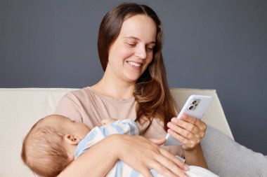 Mutlu güzel anne, akıllı telefonu internet alışverişi için kullanıyor ya da sosyal ağları kontrol ediyor küçük çocuğu kucaklıyor ve internete kayıyor.