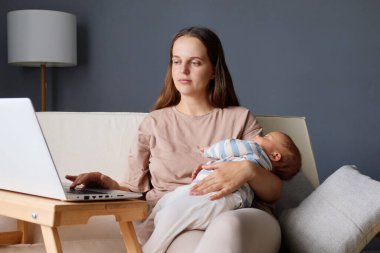 Odaklanmış bir kadın, iş ve annelik işlerini dengelemek için dizüstü bilgisayarında otururken, anne ve babasının, doğum izninde, rahat çalışma odasında uzaktan çalışan tatlı bebeği ile birlikte.