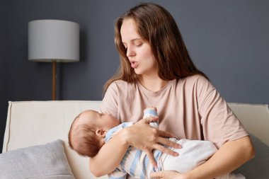 Kahverengi saçlı, sevgi dolu bir anne yeni doğmuş bebeğini elinde tutarken kanepede yavaşça sallanıyor ve küçük çocuğunu kucaklıyordu.