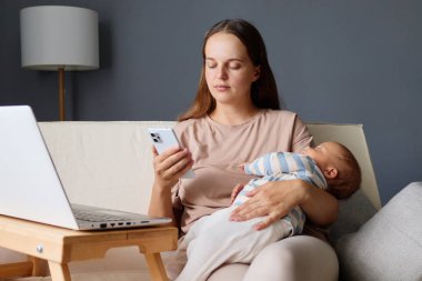 Kanepede bebeğiyle oturan odaklanmış bir kadın işe bağlı kalmak için akıllı telefonunu kullanırken evden sakince e-postaları ve sosyal medyayı karıştırıyor.