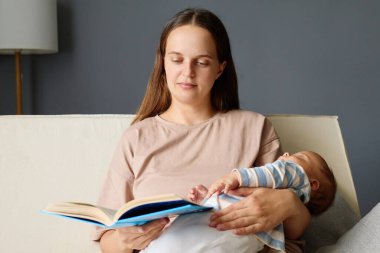 Kahverengi saçlı, konsantre bir kadın, bebeğini kucağında tutarken kanepede kitap okurken çocuğuyla vakit geçirir ve annelik dönemindeki ilginç edebiyatının tadını çıkarır.