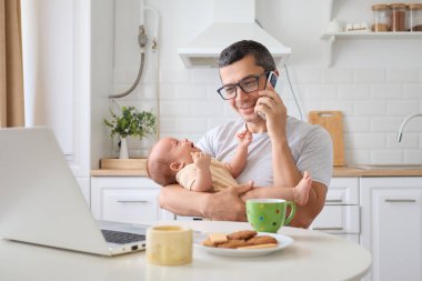 Mutlu bir baba cep telefonuyla konuşuyor. Yeni doğmuş oğlunu kucağında tutuyor. Ev mutfağında dizüstü bilgisayarında çalışıyor.