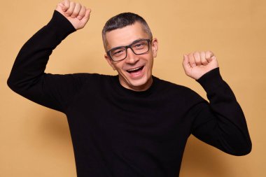 Beyaz gözlüklü ve siyah paçavralı bir adam içeride neşeli bir dansla kutlama yapıyor ve kollarını kaldırıp heyecan ve eğlence ifade ederken bej arka planda zafer kazanmış bir şekilde sırıtıyor.