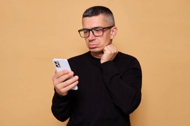 Gözlüklü ve kazaklı üzgün bir adam telefonuyla mesajları okuyor ve bej arka planda izole edilmiş üzüntü duygusunu internete aktarıyor.