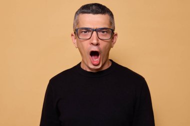 Şok olmuş gözlüklü ve siyah kazaklı bir adam kızgın görünürken rahatsız ve hayal kırıklığına uğramış bir halde neden bej arka planda soyutlandığını anlamaya çalışıyor.