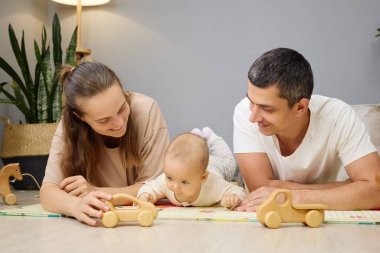 Anne ve baba yerde bebekle oynayan bir çocukla oynuyor oturma odasında ailece yerde oynuyorlar ve yeni doğmuş ebeveynler evde bebekle vakit geçiriyorlar.