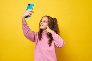 Küçük bir kız video kaydediyor ya da cep telefonuyla sosyal medya için selfie çekiyor. Çevrimiçi izleyicileriyle neşeli bir ruh hali paylaşmak için heyecanlı. Sarı arka planda izole edilmiş kalp hareketi gösteriyor.