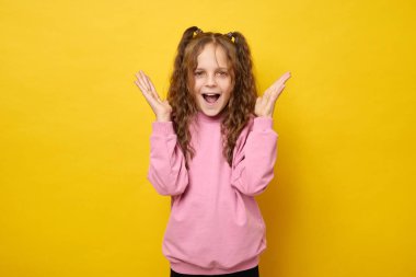 Dalgalı saçlı, şık at kuyruklu ve makyajlı küçük kız kollarını kaldırdı. Mutlu ve şaşkın bir şekilde sarı arka planda izole edilmiş mutlu bir ifadeyle haykırıyordu.