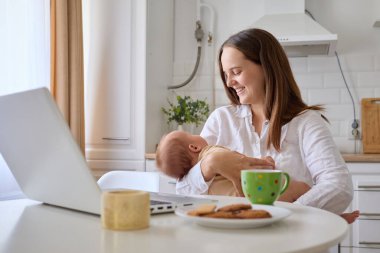 Woman smiling happily while working remotely from her kitchen on laptop with her newborn baby in hands doing her online job on her computer